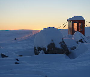 Foreground to horizen snow covered rocky ground, left middle of picture is a cabin of a tractor converted into an outhouse, sun is setting leaving an orange glow at horizen