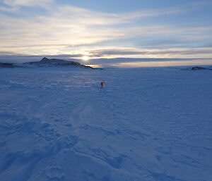 Person if fluro orange dry suit in middle of sea ice channel between Bailey Peninsula and Shirley Island, in distance rocky outcrops of island and sun is low on horizen lighting the cirrostratus cloud across the sky