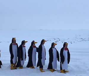 Group of seven people in penguin costumes lined up on snowy ground with white snowy background and cloud covered sky. One penguin at end of the row is facing the wrong direction