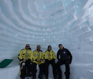Four men sitting on an ice bench inside a large igloo built of large ice blocks with blue light shining through the joins between blocks