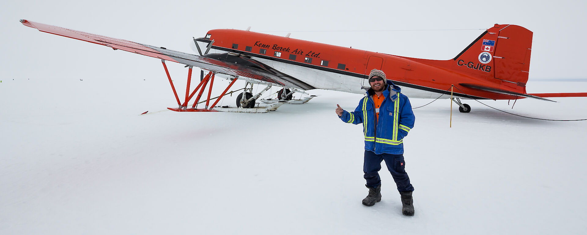 In foreground man standing smiling giving a thumbs up, in background Basler aircraft on ice runway, basler is white bottom half and red to half
