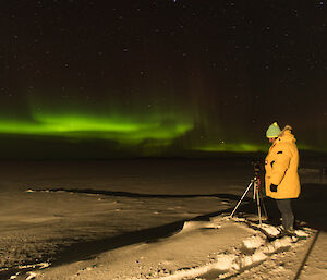 Foreground man stands on snow covered ground with camera on tripod, background night time sky with green aurora band along the horizen