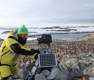 In foreground man working on a camera standing on tripod on rocky outcrop with small solar panel in front, in the background is a large Adelie penguin colony over looking a bay and snow covered hills at horizen
