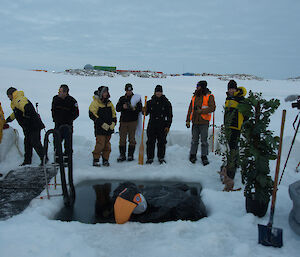 People in winter gear standing around the hole cut in the ice, Ladder is suspended into the hole. Blow up toucan is floating in the water. In the background is Casey station