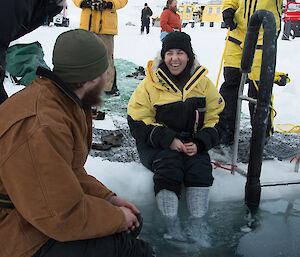 Two people sitting on the edge of the ice hole dangling their feet in the water
