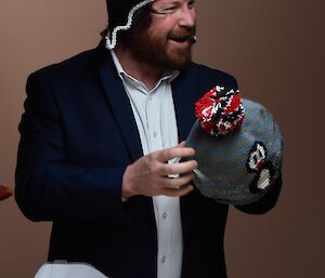 Man in suit jacket with penguin face beanie on and holding beanie with red,black and white pompom, smiling