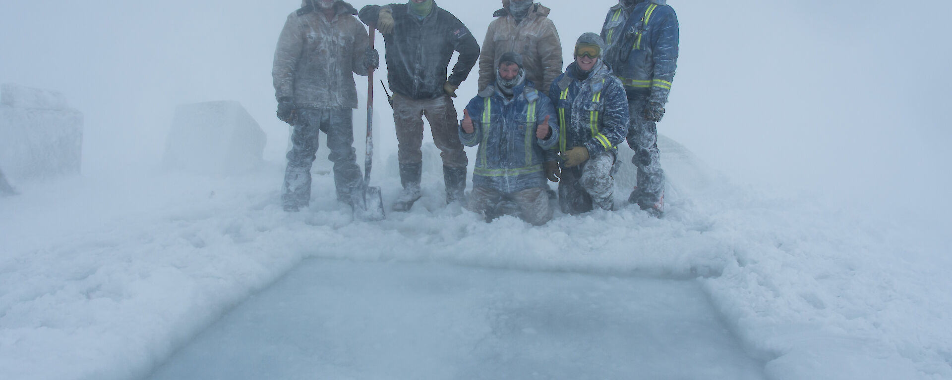 Group of men standing behind a large square hole cut in ice, they are covered in snow and the weather is white-out