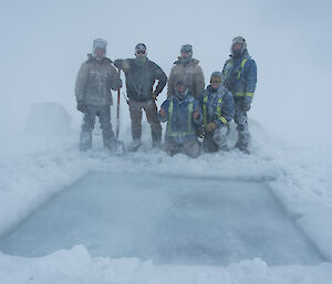 Group of men standing behind a large square hole cut in ice, they are covered in snow and the weather is white-out