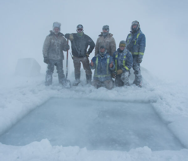 Group of men standing behind a large square hole cut in ice, they are covered in snow and the weather is white-out