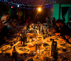 Long table laden with food and drinks, lit by fairy lights and candlelight, with expeditioners in suits and evening wear sitting at edges eating and enjoying themselves