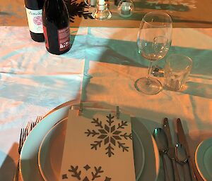 Formal place setting with crockery, cutlery and glassware with menu card on top which is cream with silver stencilled snowflakes, centre of the table has a table runner with the same stencilling pattern