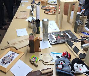 Large table covered with an array of homemade gifts including framed cross-stitch, homemade knife, beanies, bread board, wooden stool, metal photo frame