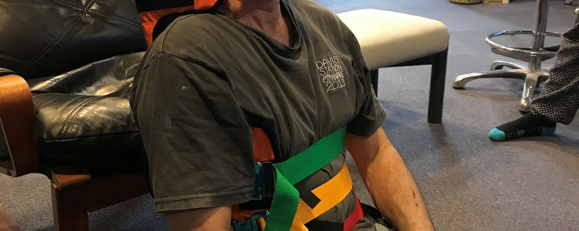 Man sitting on carpeted floor with back resting against black leather chair, strapped into back and neck brace