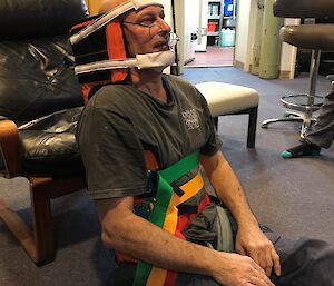 Man sitting on carpeted floor with back resting against black leather chair, strapped into back and neck brace