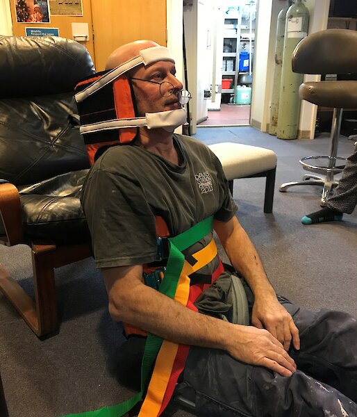 Man sitting on carpeted floor with back resting against black leather chair, strapped into back and neck brace