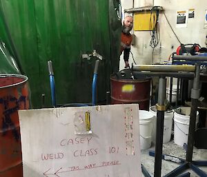 In foreground rough sign written on old cardboard “Casey Welding Class 101”, behind is metal workshop with much of the picture taken up with large green blockout curtain, but back centre a man looking out from behind curtin in high vis