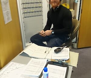 Man sits at fire alarm panel with table infront covered in paperwork