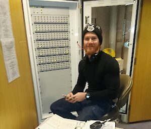 Man sits at fire alarm panel with table infront covered in paperwork