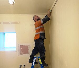 Man standing on short ladder in corner of room is sanding the walls