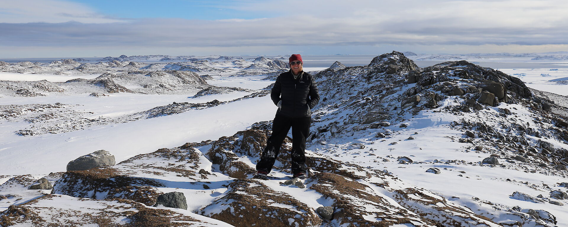 Woman in black pants and jacket with beanie and sunglasses stands centre picture, on rocky outcrop with snow covered mountainous landscape behind