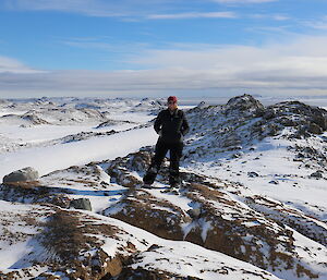 Woman in black pants and jacket with beanie and sunglasses stands centre picture, on rocky outcrop with snow covered mountainous landscape behind