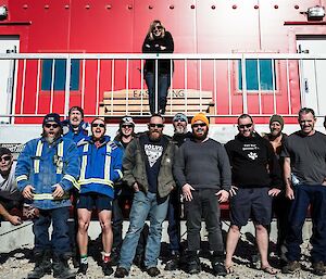 Group of expeditioners standing in foreground, one woman standing on verandah of redshed behind