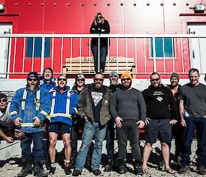 Group of expeditioners standing in foreground, one woman standing on verandah of redshed behind