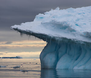 Right foreground end of large iceberg with ‘muffin top’ and icicles off edge. Left side is calm sea with sunset in background