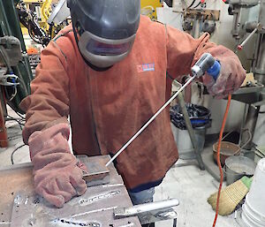 Woman in welding PPE, visor, leather jerkin and leather gloves, stands a work bench holding two small peices of metal in one hand and welding stick in other