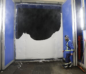Photo taken from inside of balloon shed, with blue walls and large double height doors opened. Opening is covered half way up with bank of snow. Man stands in front of door opening in cold weather gear
