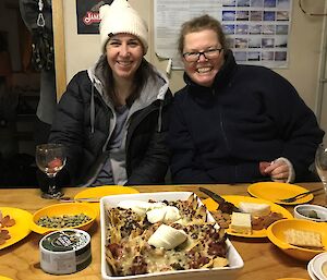 Two women sitting together behind a table laden with food which includes a big plate of nachos and a cheese platter