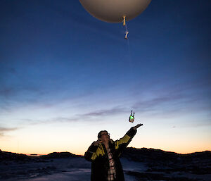 Foreground, lady in large overcoat and beanie releases large which weather balloon, in the background the sun sets over the horizen