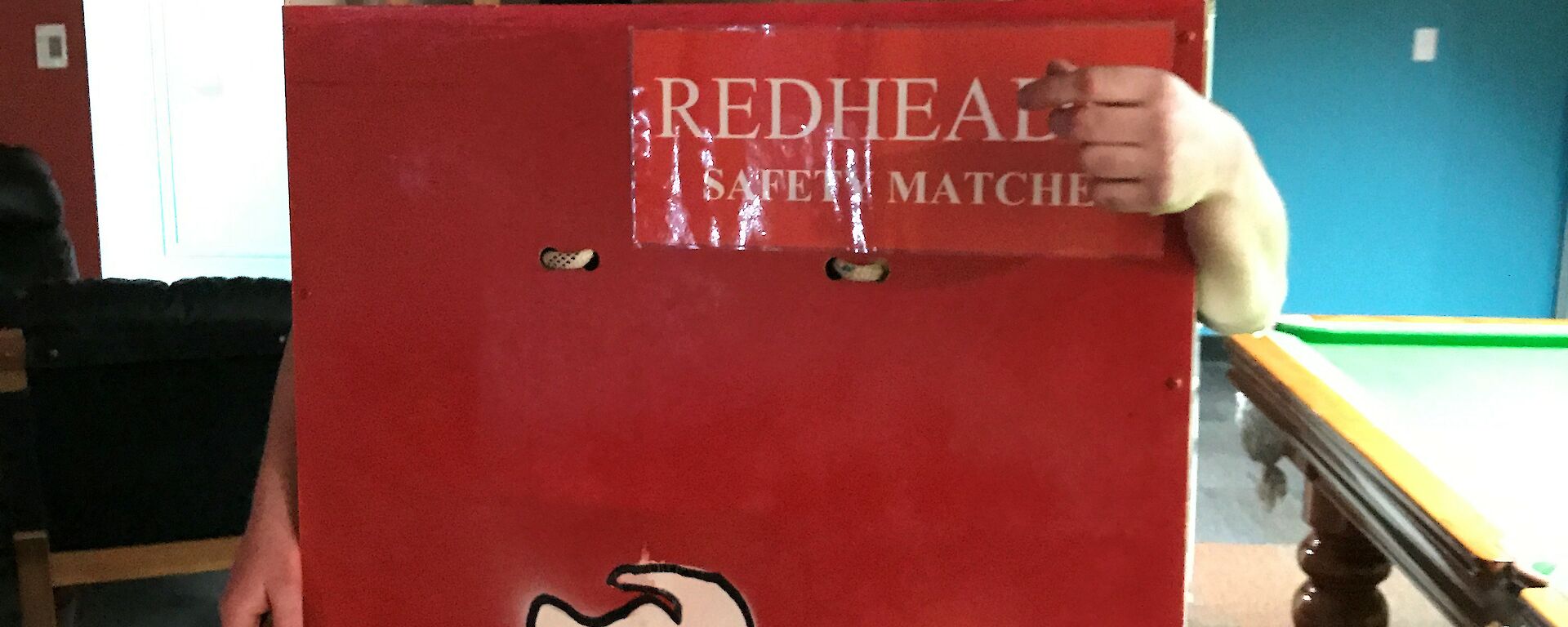 Man in box painted as redheads matches box, his face is painted red to look like the top of a match and there are large wooded matches hanging in the box beside him