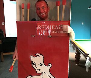 Man in box painted as redheads matches box, his face is painted red to look like the top of a match and there are large wooded matches hanging in the box beside him