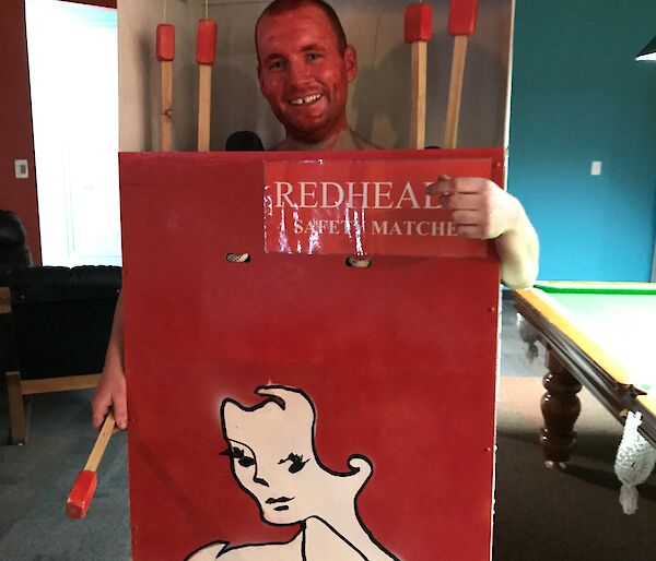 Man in box painted as redheads matches box, his face is painted red to look like the top of a match and there are large wooded matches hanging in the box beside him