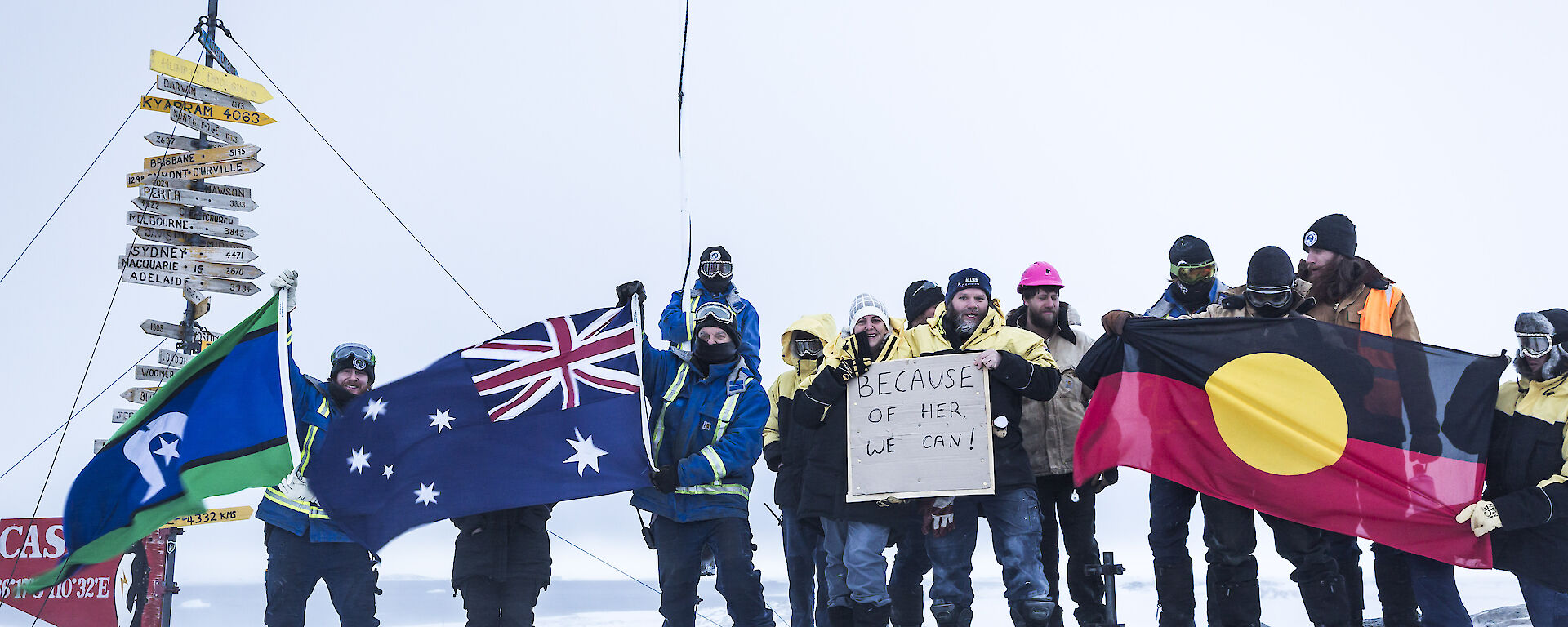 Group of expeditioners standing on rise in front of Casey sign holding the Torres Strait, Australian and Aboriginal flags and a sign reading “because of her we can”