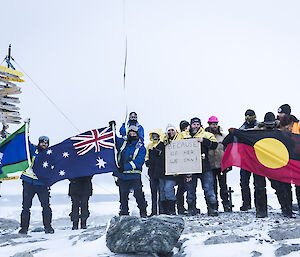 Group of expeditioners standing on rise in front of Casey sign holding the Torres Strait, Australian and Aboriginal flags and a sign reading “because of her we can”