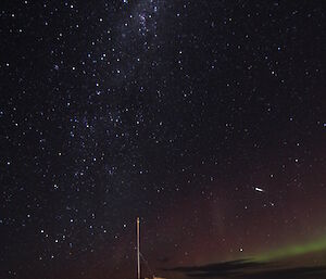 In the foreground a small wooden hut on rocky outcrop, on the horizen a pale green and red aurora, and above the expanse of the milky way with a small shooting star