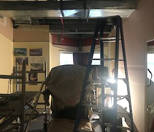 Small room filled with cardio equipment covered by drop cloths with ladder in foreground and ceiling tiles removed