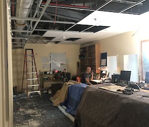 Large long room as building site, centre bench covered with drop cloths, ceiling tiles romoved, rubbish and building rubble on floor, ladder in back corner. Man sits as long desk on phone, working amongst the confusion