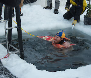 Man swimming in square hole cut in sea-ice, ladder from ice edge into the water, man is wearing a stripey ball cap and costume in the shape of a pizza slice
