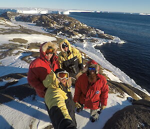 Group of four men in goose down jackets and goggles, standing on peninsula with sea and glacier in background