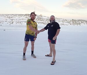 Two men in short and tshirts standing out in the snow, shaking hands. One on left is wearing wallabies shirt and on right the all blacks shirt