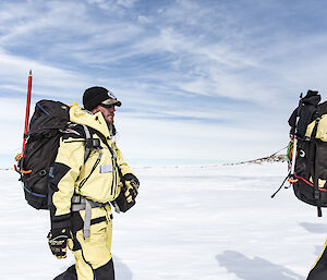 Snow to horizen with blue sky above. In foreground man is man in AAD winter uniform — yellow and black gortex jacket and pant — carrying pack