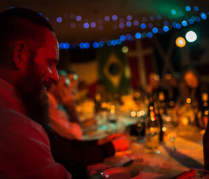 Man in forground sitting at corner of long festive table covered with crockery, glasses and bottles, with fairy lights above