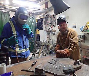 Two men standing at steel work bench, man on left in welding helmet and holding welding wand in gloved hand, man on right in leather coat and gloves with helmet on head but up