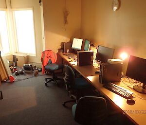 Bank of three computers with drives, keyboards and montiors along long bench from right front to centre back, with chairs at desks, and carpeted floor on left
