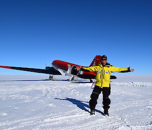 Man in AAD winter gear (yellow and black) standing in front of basler aircraft on ice runway with arms wide open. Clear blue sky above