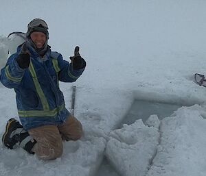 Man sits on left side a half cut out hole in ice in cold weather gear including beanie and goggles, giving two thumbs up and smiling to camera
