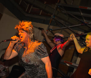 Man dressed to look like Tina Turner (spikey wig and sparkley dress) sings into microphone, with two men also dressed as ladises as back up singers